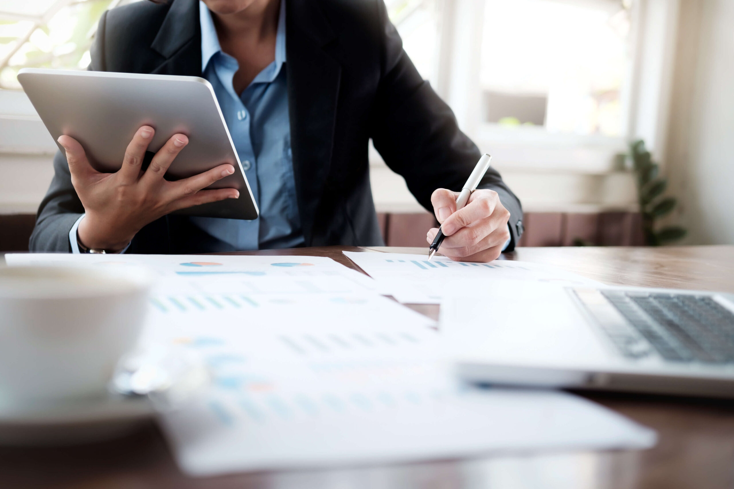 Timmins Financial A professional in business attire reviews charts and writes on documents at a desk with a laptop and tablet, showcasing Timmins Financial’s commitment to precision in a bright office setting.