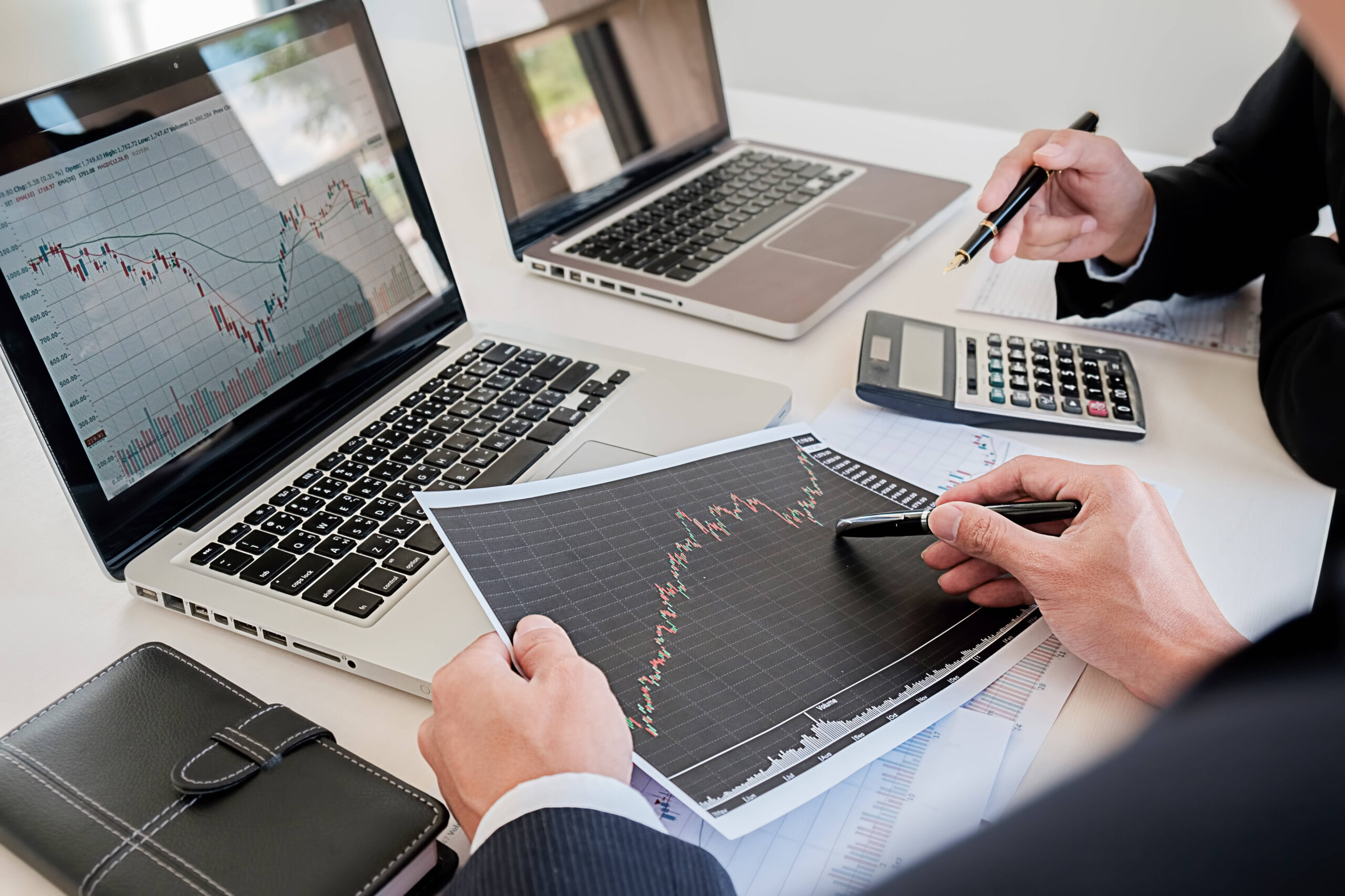 Timmins Financial Two people at a desk review a printed stock market chart, analyzing financial data displayed on their laptops. The Timmins Financial logo is visible on the screen as they discuss trends using a calculator and notebook.