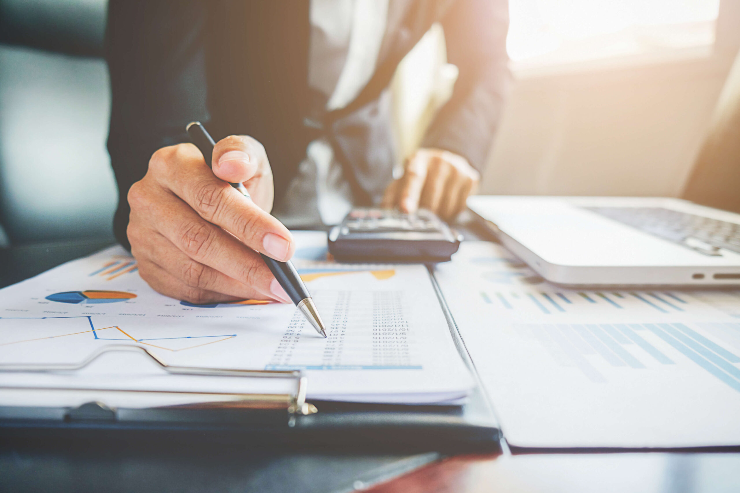 Timmins Financial A person from Timmins Financial analyzes financial charts and data on paper, using a pen, calculator, and laptop at their office desk.