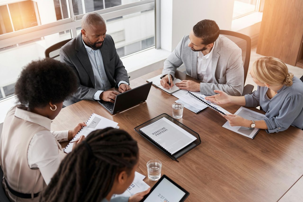 Timmins Financial Five people in business attire sit around a table with documents and laptops, engaged in a Timmins Financial meeting in a bright, modern office.