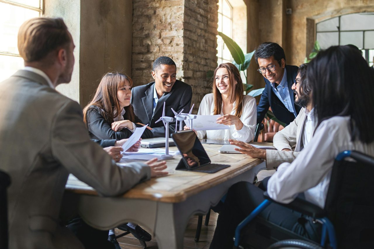 Timmins Financial A diverse group of professionals from Timmins Financial sit around a table in a modern office, discussing documents and smiling during a meeting.