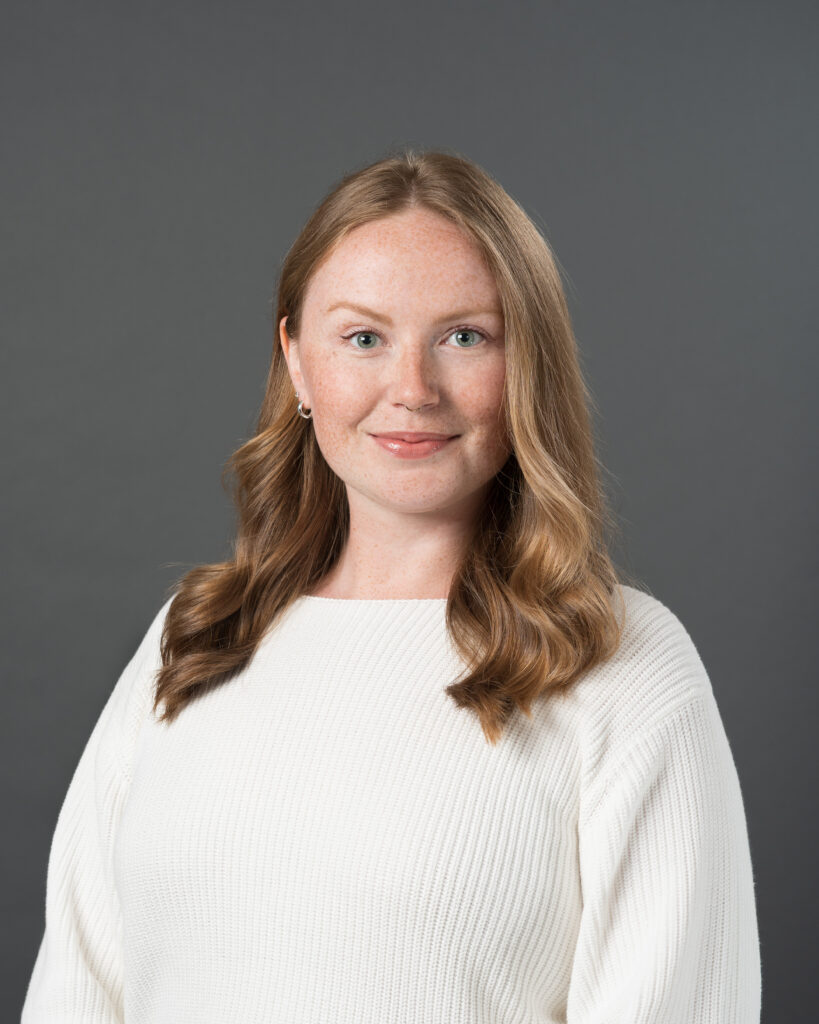 Timmins Financial A woman with long, wavy, light brown hair and fair skin, wearing a white sweater, poses against a plain gray background in a Timmins Financial promotional shoot.