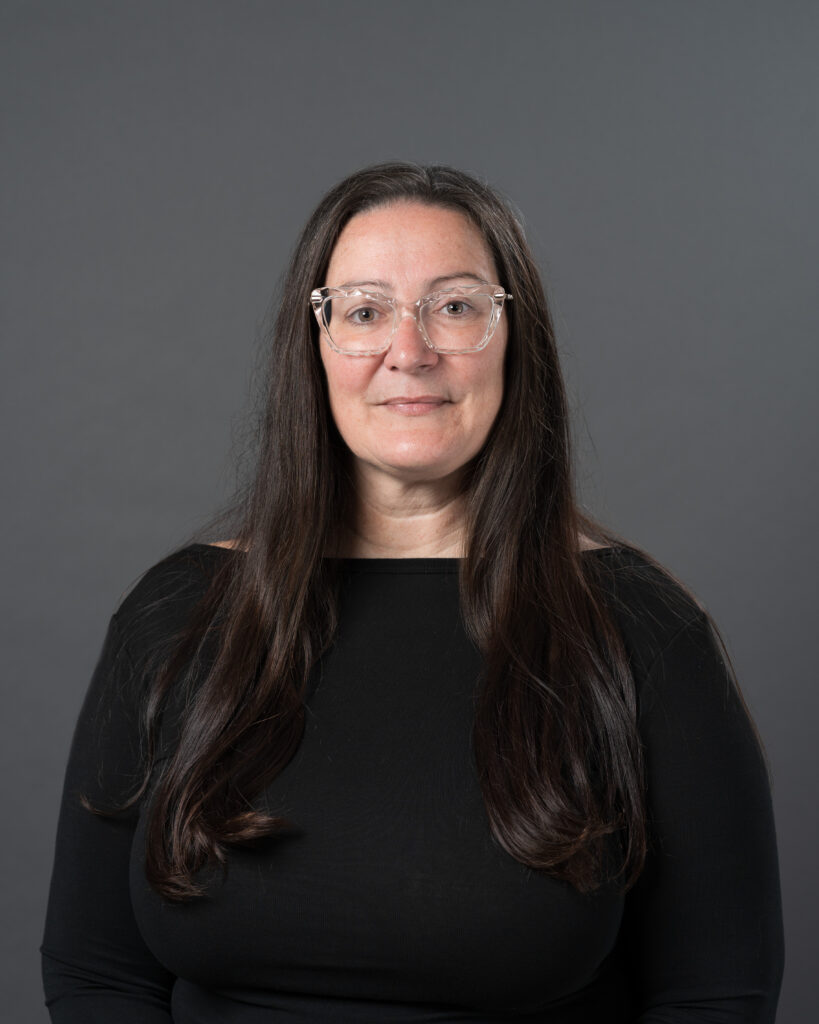 Timmins Financial A woman with long brown hair, wearing clear glasses and a black top, poses confidently in front of a plain dark gray background, reflecting the professional image of Timmins Financial.