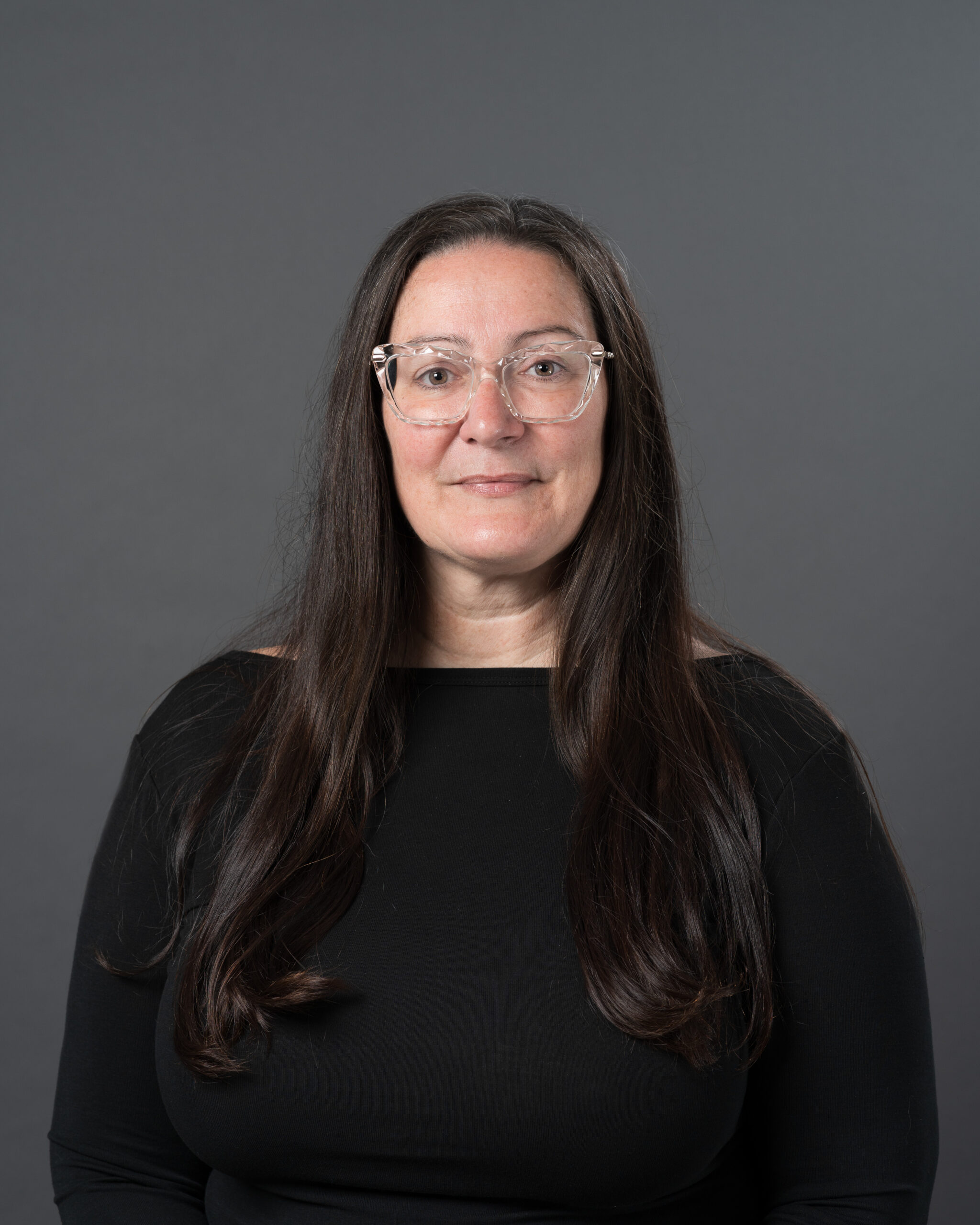 Timmins Financial A woman with long brown hair, wearing clear glasses and a black top, poses confidently in front of a plain dark gray background, reflecting the professional image of Timmins Financial.