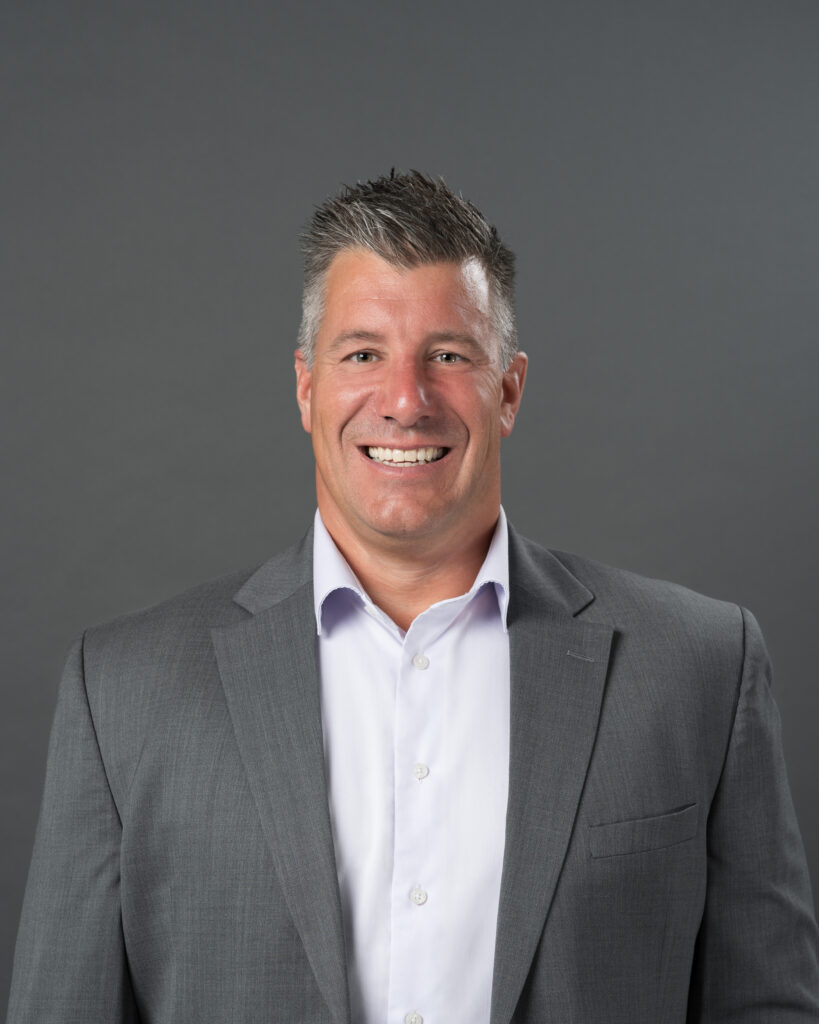 Timmins Financial A man in a gray suit and white shirt, representing Timmins Financial, smiles at the camera against a plain gray background.