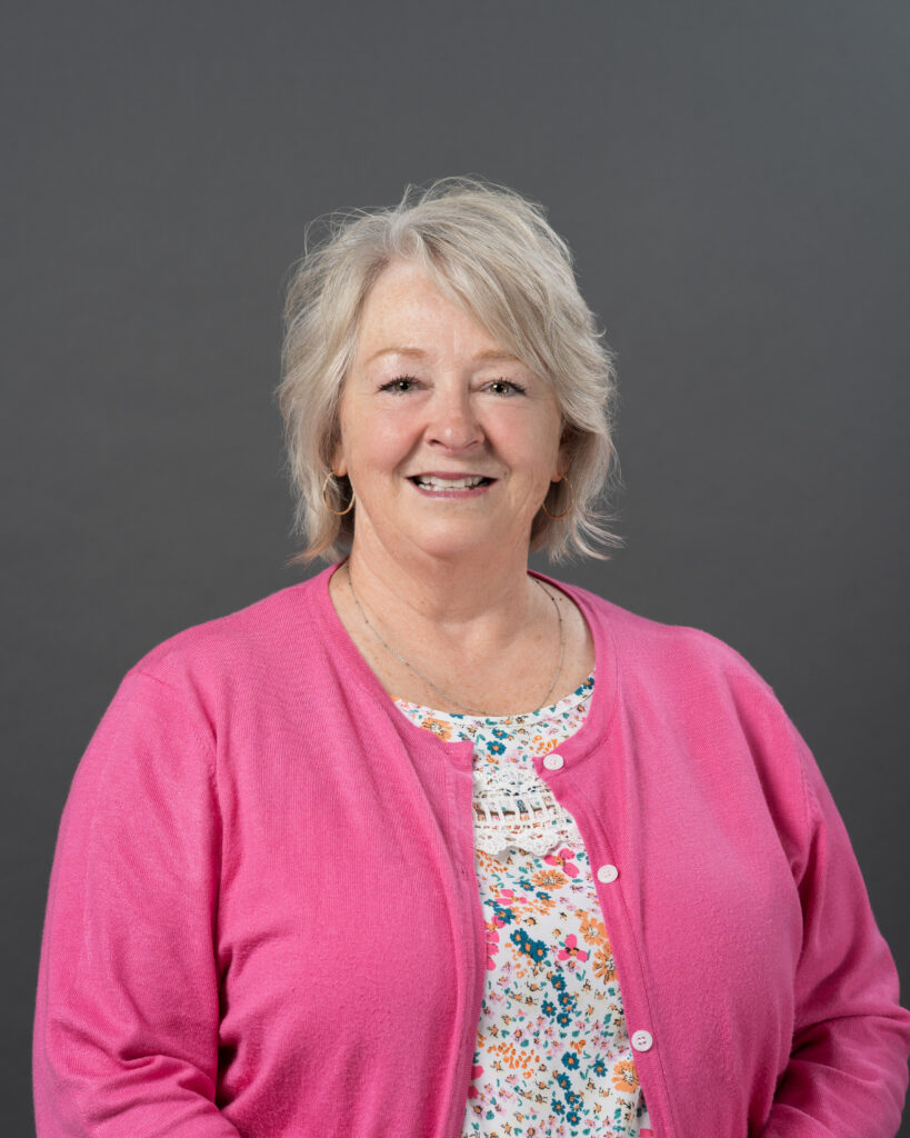 Timmins Financial Older woman with short gray hair, wearing a pink cardigan over a floral top, smiles warmly in front of a plain gray background, reflecting the trust and confidence valued by Timmins Financial.