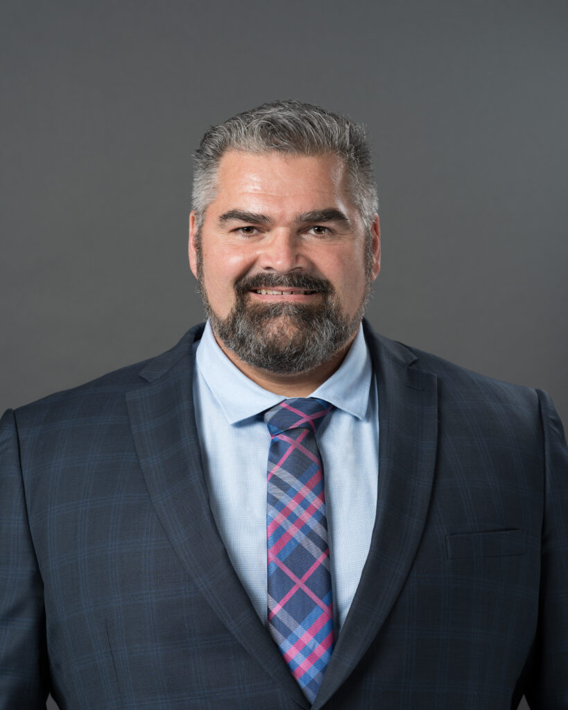 Timmins Financial Man with short gray hair and beard wearing a navy plaid suit jacket, light blue shirt, and pink and blue plaid tie, posing against a plain gray background for Timmins Financial.