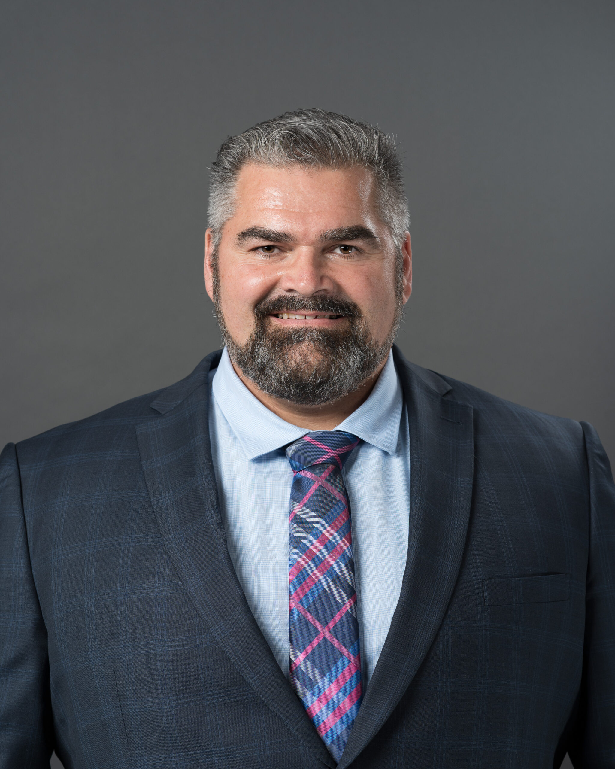 Timmins Financial Man with short gray hair and beard wearing a navy plaid suit jacket, light blue shirt, and pink and blue plaid tie, posing against a plain gray background for Timmins Financial.