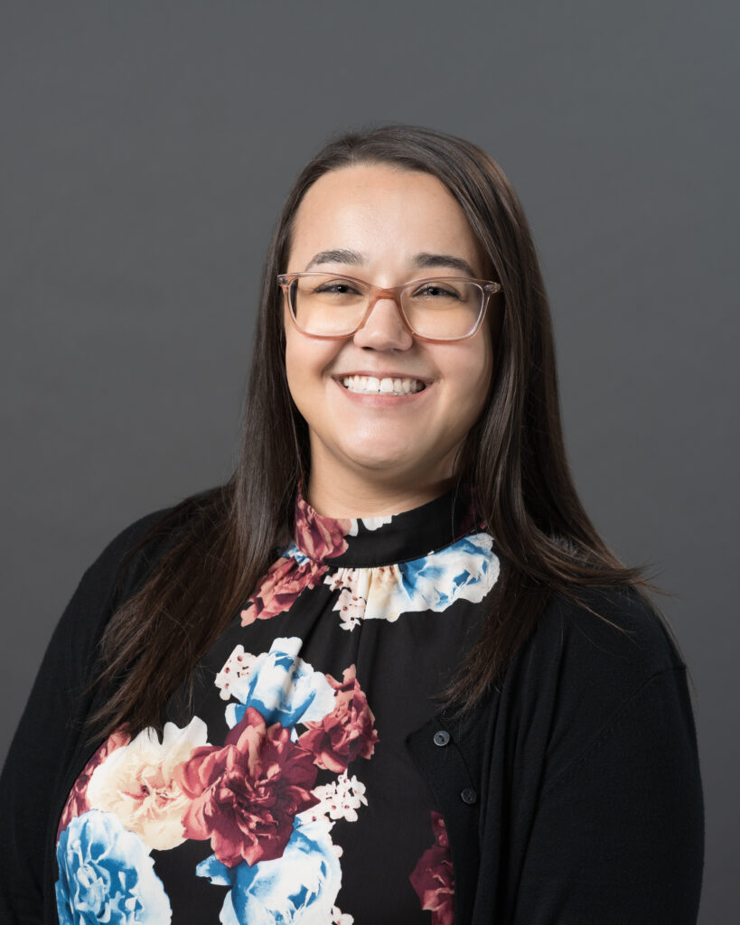 Timmins Financial A woman with long brown hair and glasses smiles at the camera, wearing a black floral top and black cardigan, representing Timmins Financial against a plain gray background.