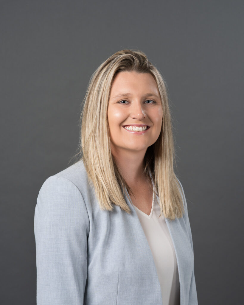 Timmins Financial A woman with straight blonde hair, wearing a light gray blazer and white blouse, poses in front of a plain gray background, smiling at the camera—her polished look reflects the professionalism of Timmins Financial.