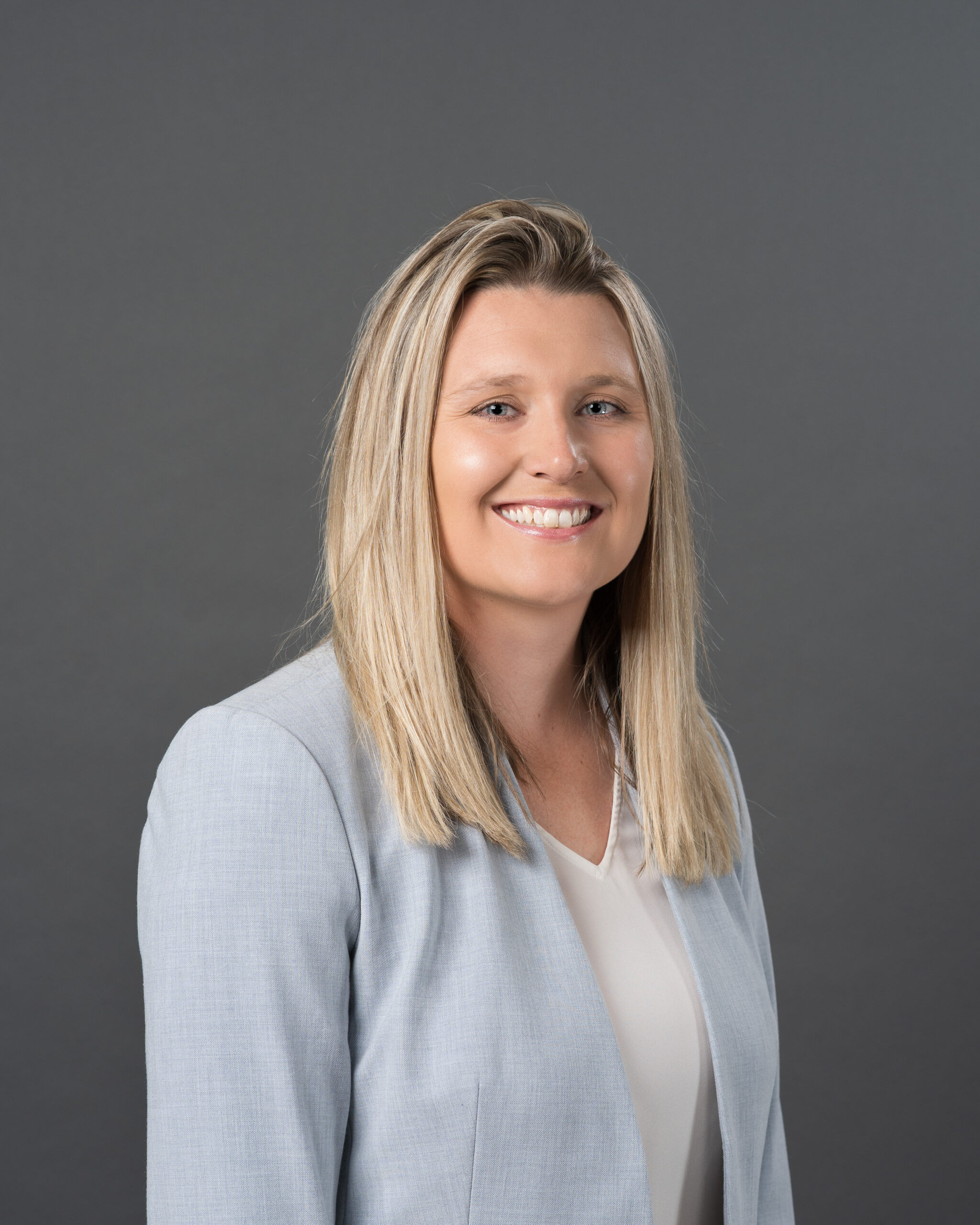 Timmins Financial A woman with long blonde hair wearing a light gray blazer and white blouse poses in front of a plain gray background, smiling confidently at the camera—a professional look perfect for Timmins Financial.