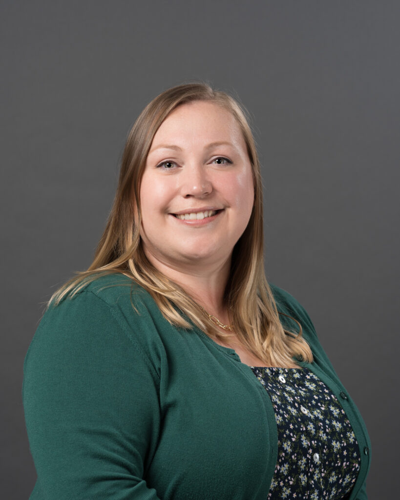 Timmins Financial A woman with long blonde hair, wearing a green cardigan and a floral top, smiles at the camera against a plain gray background, reflecting the friendly and approachable spirit of Timmins Financial.