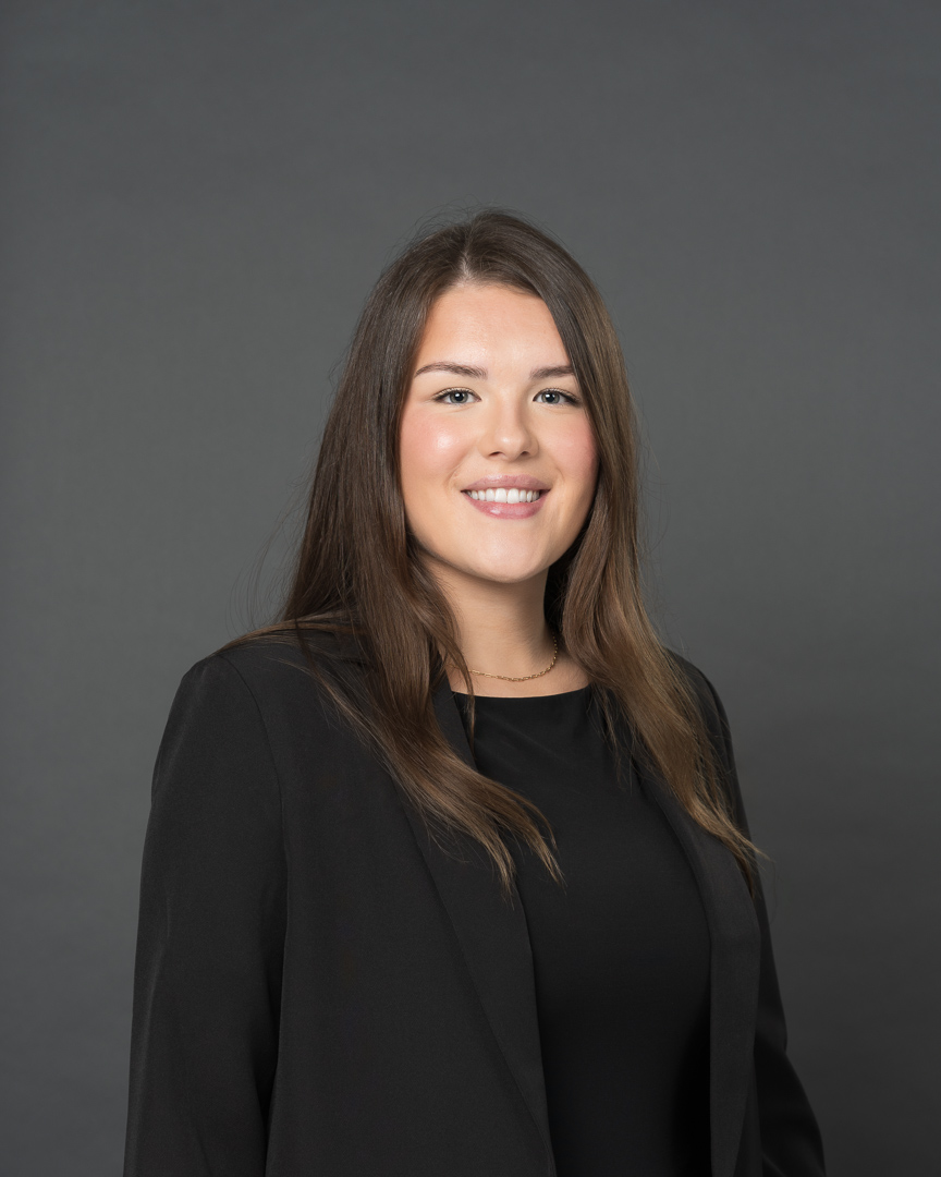 Timmins Financial A woman with long brown hair, wearing a black blazer and top, stands against a plain gray background, facing the camera and smiling.